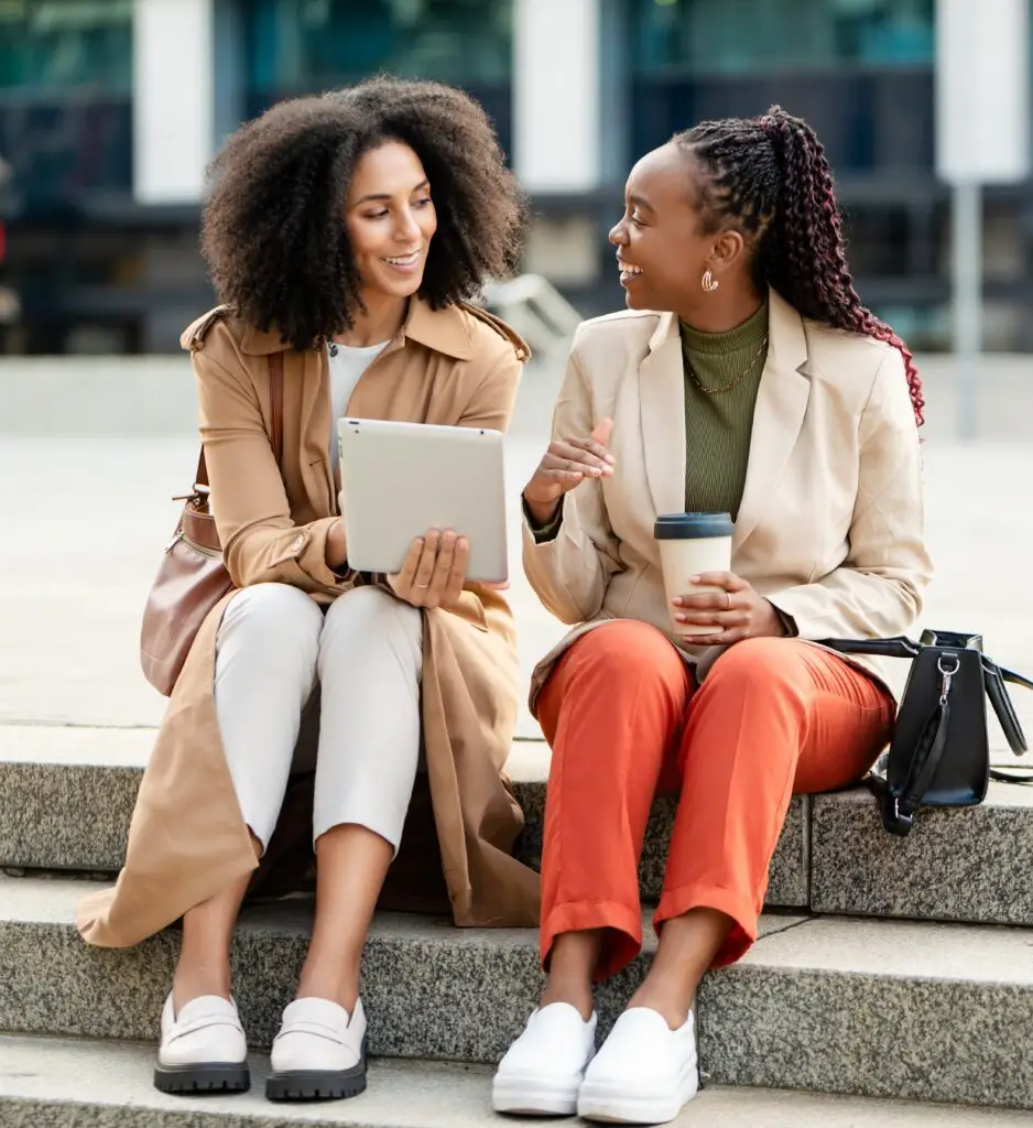 Two Black businesswomen engage in a lively discussion while seated on outdoor steps. They are reviewing information on a tablet, indicating a professional business setting. The image portrays entrepreneurship, collaboration, and teamwork.